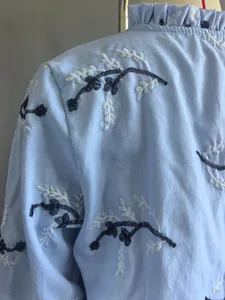 Detailopname van een lichtblauwe blouse met bloemenborduursel en rucheskraag.