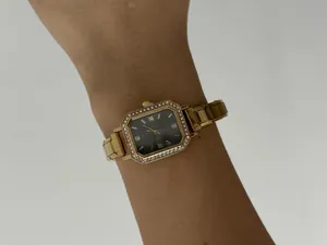 Gros plan d'une montre carrée dorée élégante avec lunette sertie de cristaux sur un poignet.