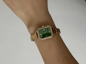 Gros plan d'une montre dorée pour femme avec cadran carré vert et lunette strass.