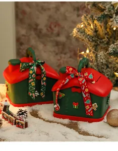 Scatole regalo di Natale a forma di casa con nastri in una scena festosa con albero e neve.