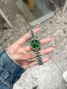 Hand hält eine Dinimi Damenuhr mit smaragdgrünem Zifferblatt und Strass-Lünette-Detail.