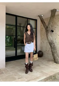 Full body shot of a woman wearing a brown V-neck knit vest and white denim mini skirt.