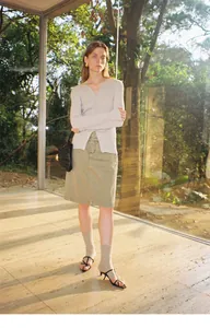 Model met lichtbeige gebreid vest met V-hals en olijfgroene rok in een tuinsetting