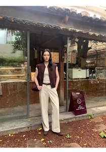 Modella asiatica con gilet di camoscio marrone Maillard e pantaloni beige davanti a un caffè.