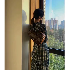 Mujer con vestido de cuadros y mochila de cuero marrón vintage junto a una ventana al atardecer.