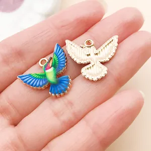 Détail rapproché de pendentifs colibris en émail coloré et ton or tenus à la main