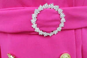 Dettaglio di un abito blazer rosa con fibbia di strass e bottone dorato decorativo.