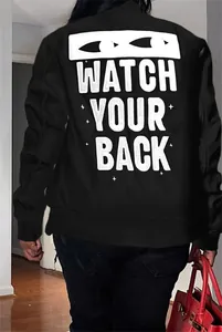 Detail view of black bomber jacket back with white Watch Your Back slogan print.