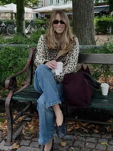 Stilvolle Frau in Leopardenjacke genießt einen Kaffee auf einer Bank im Park.