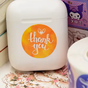 Vista detallada de una pegatina redonda naranja 'Thank You' con corazón en estuche