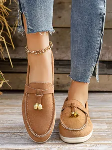 Close-up detail of brown suede slip-on loafers featuring gold bell charms and white stitching