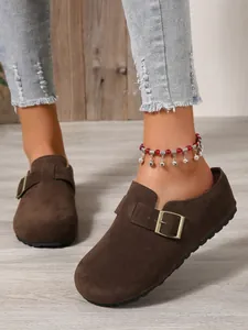 Close-up of dark brown suede slip-on mules with metal buckle and rustic background.