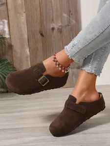 Close-up view of brown suede-like flat clogs worn with denim jeans and a red bead anklet.