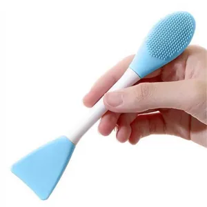 Hand mit einer zweiköpfigen blauen Silikon-Gesichtsmaskenbürste mit Spatel und Bürste.