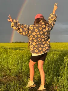 Stylish woman in a grey floral fleece sweatshirt standing in a field under a rainbow