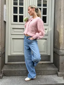 Model in süßem rosa Strickcardigan und Jeans vor einer klassischen weißen Holztür