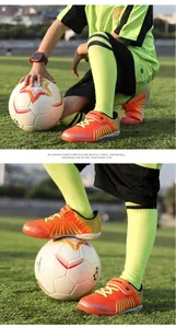 Professional detail view of orange children's TF soccer shoes on a green grass field with a ball