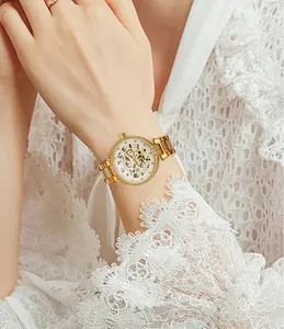 Elegante goldene Damen-Skelettuhr mit Strasssteinen vor einem weißen Spitzenärmel-Hintergrund.