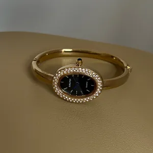 Nahaufnahme einer goldenen ovalen Armreif-Uhr mit Strass-Lünette und schwarzem Zifferblatt.