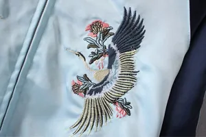 Detailaufnahme der aufwendigen Kranich-Stickerei auf einer hellblauen Satin-Bomberjacke.