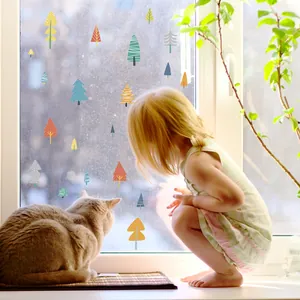 A young child and cat looking at colorful forest tree window decals on glass in a sunny room.