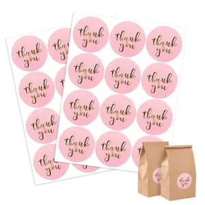 Pink round thank you stickers with gold foil cursive text shown on kraft gift bags