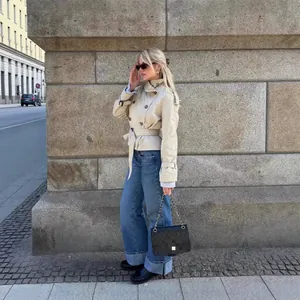 Donna elegante in trench corto beige con cintura e jeans blu in una strada urbana.
