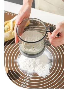 Close-up action shot of a hand-held semi-automatic flour sifter with measurement markings.