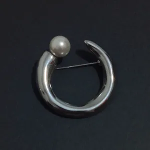 Detail of a minimalist silver metallic C-shaped brooch with a single pearl accent.