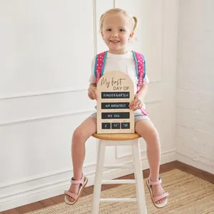 Young girl sitting on a stool holding a wooden First Day of School milestone board