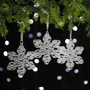 Nahaufnahme von drei transparenten Acryl-Schneeflocken-Anhängern an Tannenzweigen.