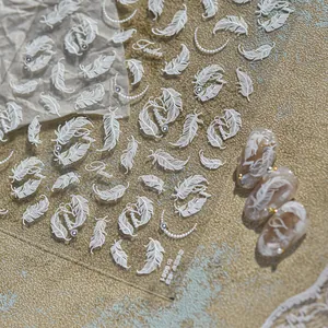Detailed view of white 3D embossed feather nail stickers on a shimmering gold surface.