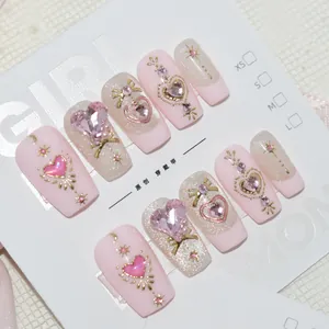 Détail de faux ongles roses faits main avec des gemmes en cœur et des nœuds dorés.