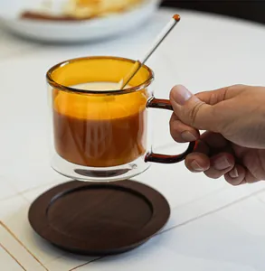 Close-up of a hand holding an amber double-walled glass coffee cup with latte and stirrer