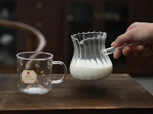 Hand holding a ribbed glass milk pitcher next to a small floral patterned glass mug.