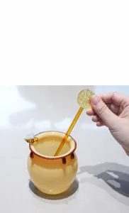 Detailed view of a honey pot shaped glass cup with a bee decoration and lemon stirrer.