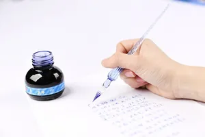 Hand writing with an elegant glass dip pen on white paper next to a blue ink bottle.