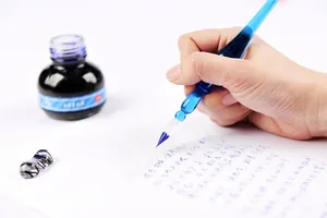Close-up detail of a blue glass dip pen writing on paper with a bottle of dark ink