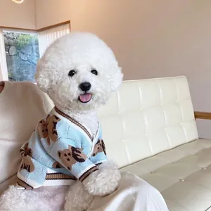 Perro blanco con cárdigan tejido azul claro y estampado de osos marrones, sentado en un sofá crema
