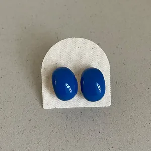 Close-up of blue glossy bean-shaped enamel stud earrings on a textured white card