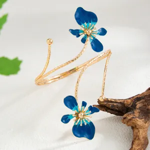 Détail d'un bracelet de bras doré avec deux fleurs en émail bleu sur fond blanc texturé.