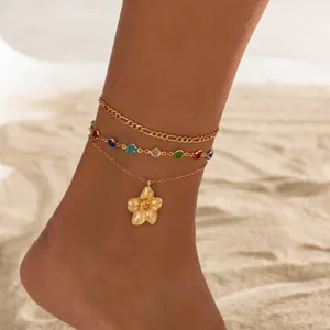 Detail shot of multilayered gold anklet with flower charm and colorful stones on sand.