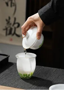 Elegant wird Tee aus einem weißen Gaiwan in eine matte Glastasse mit grünen Punkten gegossen