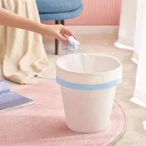 Hand throwing crumpled paper into a white trash bin with a drawstring garbage bag.