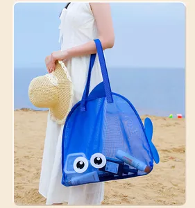 Detail view showing a person holding the blue fish-shaped mesh beach bag on a sunny beach