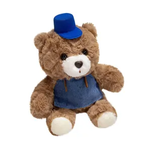 Cute brown teddy bear plush toy wearing a blue hat and denim bib on white background