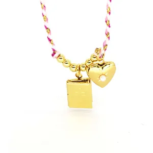 Macro detail of a gold heart and book locket pendant necklace with pink braided cord.