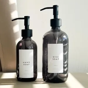 Two smoke grey glass soap dispenser bottles with labels under natural window lighting