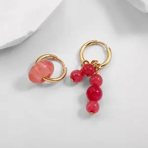 Detail of asymmetrical gold hoop earrings with red textured stone beads on a white surface.