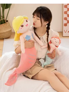 A model holding soft pink and green mermaid plush dolls in a cozy indoor setting